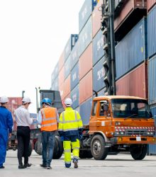 Back of professional engineer, worker or technicians walk and discuss together about work in cargo container workplace area with crane, truck and stack of container tank on background.